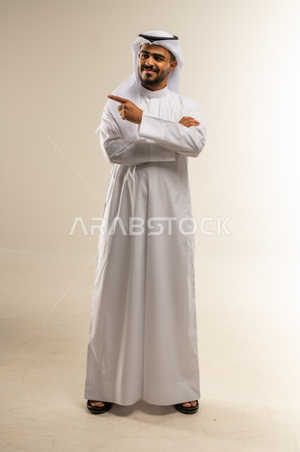 الإشارة والتوضيح وتحديد موقع معين، ايماءات السعادة والسرور، بورتريه لشاب عربي خليجي كويتي يرتدي الثوب التقليدي يشير باصبع السبابة إلى شيء ما، صورة الجسم بالطول الكامل، خلفية بيضاء