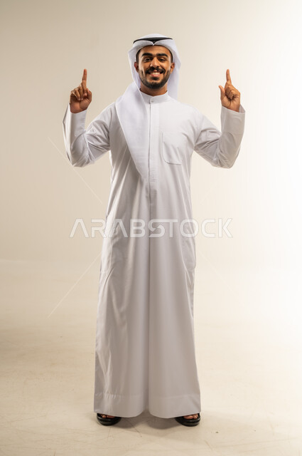 النظر الى الكاميرا بايماءات السعادة والسرور، تحديد موقع معين والإشارة والتوضيح إلى شيء ما، بورتريه لشاب عربي خليجي كويتي يرتدي الثوب التقليدي يرفع يديه واصبعه السبابة إلى الأعلى، صورة لجسم بالطول الكامل، خلفية بيضاء