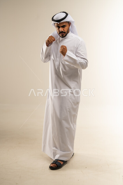 ممارسة رياضة الدفاع عن النفس، تعزيز صحة العظام والمفاصل، إتقان فنون الملاكمة، زيادة قدرة العضلات على التحمل، بورتريه لرجل عربي خليجي كويتي يرتدي الثوب التقليدي يرفع قبضات يديه بإيماءات القوة، صورة الجسم بالطول الكامل, خلفية بيضاء