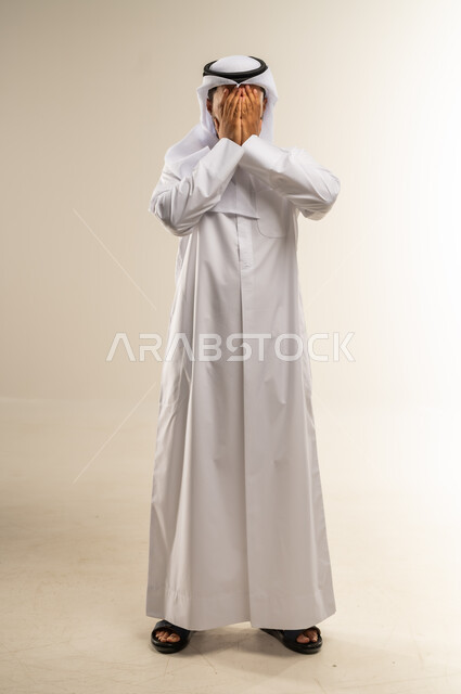 الإكثار من الدعاء في شهر رمضان المبارك، الإجتهاد في العبادة والحفاظ على الطاعات، بورتريه لرجل عربي خليجي كويتي يرتدي الثوب التقليدي يضع يديه على وجهه ويدعو الله بإيماءات التوسل والتضرع، الطول الكامل للجسم, خلفية بيضاء