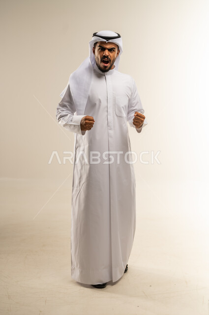 مفهوم الغضب والانفعال, بورتريه لرجل عربي خليجي كويتي يرتدي الثوب التقليدي ينظر الى الكاميرا يرفع يديه عاليا بإيماءات التهيج و التوتر, تعابير الصراخ والضغط النفسي, صورة الجسم بالطول الكامل, خلفية بيضاء