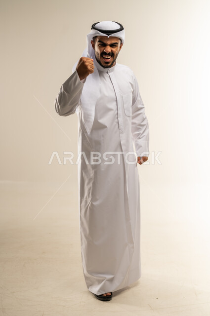 الانتصار والفوز والنجاح، مفهوم الانجاز وتحقيق الاهداف، بورتريه لشاب عربي خليجي كويتي يرتدي الثوب التقليدي يرفع قبضة يديه للاعلى بإيماءات الفرح والسرور, الشعور بالسعادة والقوة، صورة الجسم بالطول الكامل، خلفية بيضاء