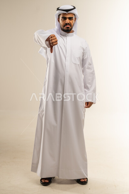القلق والفشل في تحقيق أمر ما، بورتريه لرجل عربي خليجي كويتي يرتدي الثوب التقليدي ينظر الى الكاميرا يشير بإصبع الابهام الى الاسفل بإيماءات الانزعاج والرفض، تعابير عدم القبول والرضا، الجسم بالطول الكامل، خلفية بيضاء