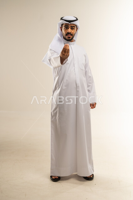 مفهوم الصبر والتريث و التأني، بورتريه لرجل عربي خليجي كويتي يرتدي الثوب التقليدي ينظر الى الكاميرا يرفع يده نحو الأعلى بإيماءات طلب الإنتظار والتمهل, إمهال بعض الوقت لفعل شيء ما، صورة الجسم بالطول الكامل، خلفية بيضاء