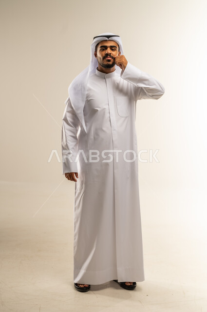 مفهوم الرجولة والقوة، التهديد والتوعد لشيء ما، بورتريه لرجل عربي خليجي كويتي يرتدي الثوب التقليدي ينظر الى الكاميرا ويمسك شاربه، إيماءات الالتزام والوفاء بالوعود، حركات وتعابير شبابيه، الطول الكامل للجسم، خلفية بيضاء
