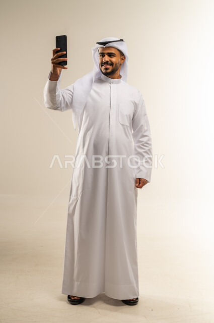 التقاط صور السيلفي التذكارية، مفهوم التقنيات والأجهزة الحديثة، بورتريه لرجل عربي خليجي كويتي مبتسم يرتدي الثوب التقليدي يحمل بيده الجوال، صورة الجسم بالطول الكامل، خلفية بيضاء