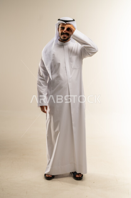 Focusing and looking at a specific thing, trying to bring distant things closer, portrait of a Kuwaiti Gulf Arab man wearing traditional dress, putting his hand over his eyes in the shape of a lens with gestures of searching and exploring, full-length body image, white background