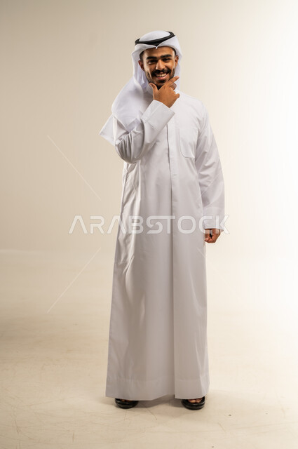 الوقوف باستقامة والنظر إلى الكاميرا بإيماءات السعادة والسرور، مفهوم الوسامة والثقة بالنفس، بورتريه لرجل عربي خليجي كويتي مبتسم يرتدي الثوب التقليدي والغتره يضع يده أسفل ذقنه، الاهتمام بالمظهر الخارجي، الجسم بالطول الكامل، خلفية بيضاء