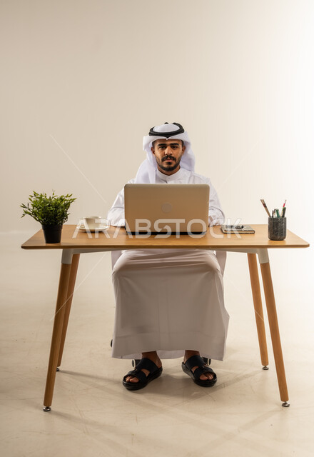 دمج التقنيات الحديثة بالأعمال الإدارية، إنجاز المهام باستخدام الحاسوب المحمول، التركيز والتفاني والعمل الجاد، التوظيف في المكاتب والشركات الكويتية، بورتريه لرجل عربي خليجي كويتي يرتدي الثوب التقليدي والغترة يجلس خلف طاولة المكتب ويعمل بجد، خلفية بيضاء