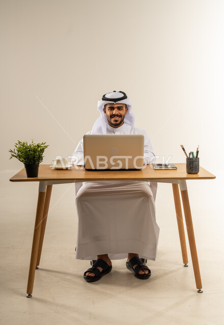 دمج التقنيات الحديثة بالأعمال الإدارية، إنجاز المهام باستخدام الحاسوب المحمول، التركيز والتفاني والعمل الجاد، التوظيف في المكاتب والشركات الكويتية، بورتريه لرجل عربي خليجي كويتي يرتدي الثوب التقليدي والغترة يجلس خلف طاولة المكتب ويعمل بجد، خلفية بيضاء