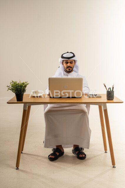 إنجاز المهام باستخدام الحاسوب المحمول، دمج التقنيات الحديثة بالأعمال الإدارية، التركيز والتفاني والعمل الجاد، التوظيف في المكاتب والشركات الكويتية، بورتريه لرجل عربي خليجي كويتي يرتدي الثوب التقليدي يجلس خلف طاولة المكتب ويعمل بجد، خلفية بيضاء