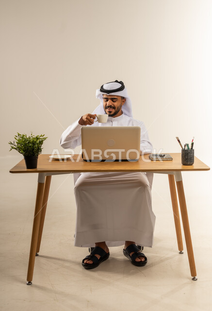 شرب المشروبات الساخنة اثناء العمل، إنجاز المهام باستخدام الحاسوب المحمول، دمج التقنيات الحديثة بالأعمال الإدارية، التوظيف في المكاتب والشركات الكويتية، بورتريه لرجل عربي خليجي كويتي يرتدي الثوب التقليدي يجلس خلف طاولة المكتب ويعمل بجد، خلفية بيضاء