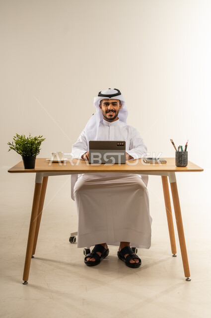 النظر الى الكاميرا بتعابير السرور، استخدام التقنيات الحديثة والمتطورة في العمل، مفهوم ادارة الاعمال عن بعد, بورتريه لرجل عربي خليجي كويتي يرتدي الثوب التقليدي يجلس خلف طاولة خشبية يحمل بيديه جهاز التابليت بايماءات السعادة, خلفية بيضاء