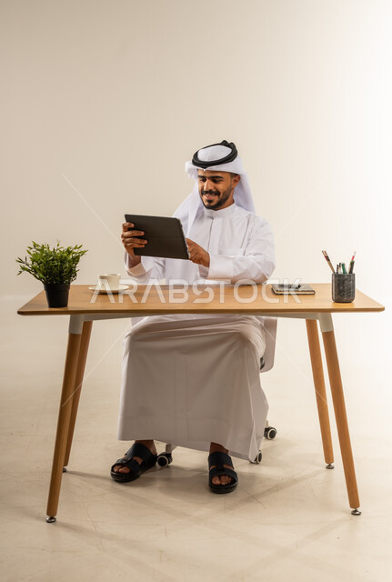 Using modern and advanced technologies in work, the concept of remote business management, portrait of a Kuwaiti Gulf Arab man wearing traditional clothing sitting behind a wooden table holding a tablet in his hands with gestures of happiness, white background