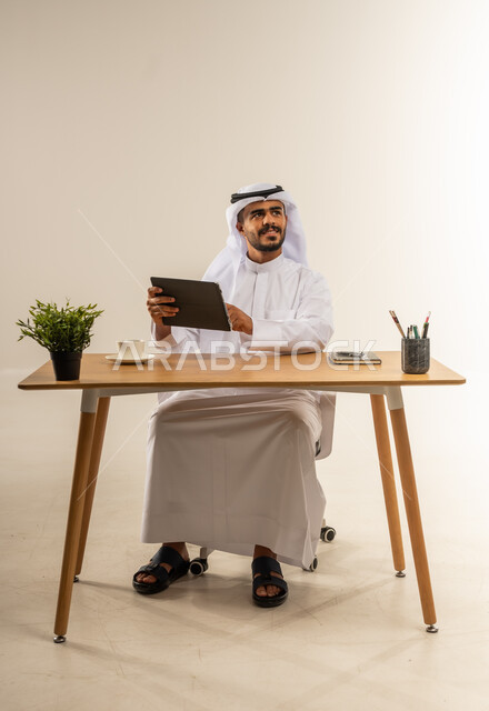مفهوم ادارة الاعمال عن بعد, استخدام التقنيات الحديثة والمتطورة في العمل، بورتريه لرجل عربي خليجي كويتي يرتدي الثوب التقليدي يجلس خلف طاولة خشبية يحمل بيديه جهاز التابليت ينظر الى شيء ما بايماءات السعادة, خلفية بيضاء