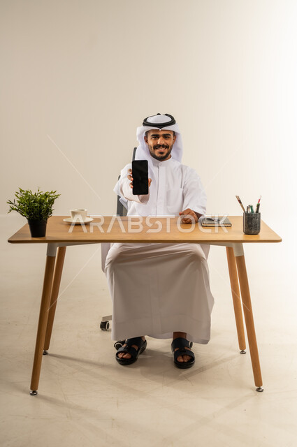 عرض شاشة سوداء فارغة، تصفح الانترنت والتسوق الالكتروني, مفهوم التقنيات والأجهزة الحديثة، استخدام الهاتف المحمول في التواصل مع الأفراد، بورتريه لرجل عربي خليجي كويتي مبتسم يرتدي الثوب التقليدي يجلس خلف طاولة خشبية يحمل بيده الجوال، خلفية بيضاء
