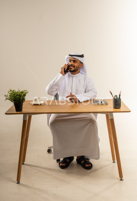 إجراء مكالمات العمل الهاتفية عبر الجوال، دمج التقنيات الحديثة بالمجال الإداري والمكتبي، التوظيف في المكاتب الكويتية، بورتريه لرجل عربي خليجي كويتي يرتدي الثوب التقليدي يجلس خلف المكتب بجانبه نبتة خضراء ويقوم بالرد والإجابة على الاتصالات، خلفية بيضاء
