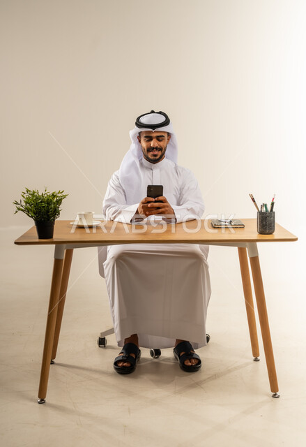 Browsing the internet and online shopping, using a mobile phone to communicate with people, portrait of a smiling Kuwaiti Gulf Arab man wearing traditional dress sitting behind a wooden table holding a mobile phone in his hand, white background