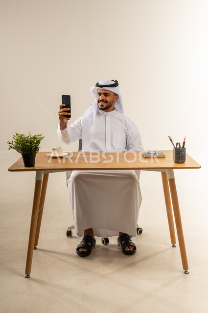 تصفح الانترنت والتسوق الالكتروني، استخدام الهاتف المحمول في التواصل مع الأفراد، بورتريه لرجل عربي خليجي كويتي مبتسم يرتدي الثوب التقليدي يجلس خلف طاولة خشبية يحمل بيده الجوال، خلفية بيضاء