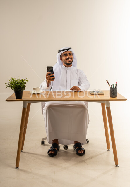 Integrating modern technologies into the administrative and office field, working in Kuwaiti offices and companies, enjoying a cup of hot coffee, portrait of a Kuwaiti Gulf Arab man wearing traditional thobe sitting behind a desk next to a green plant and holding a mobile phone, white background