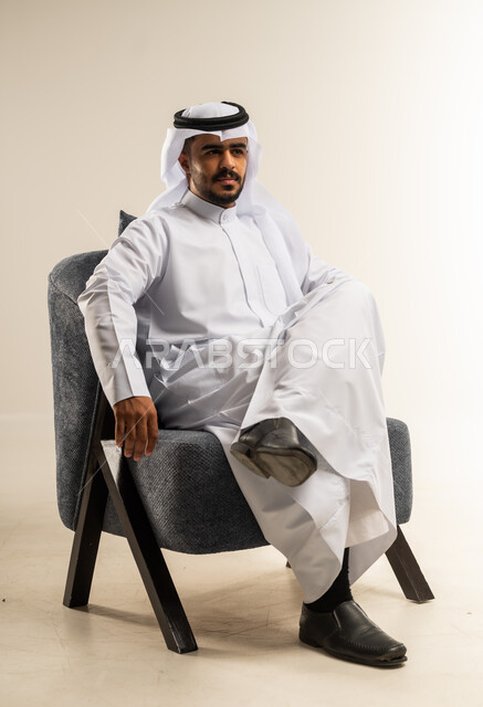 Concern for appearance and elegance, the concept of masculinity and self-confidence, portrait of a Kuwaiti Gulf Arab man wearing traditional thobe sitting on a comfortable chair with one leg crossed over the other, white background