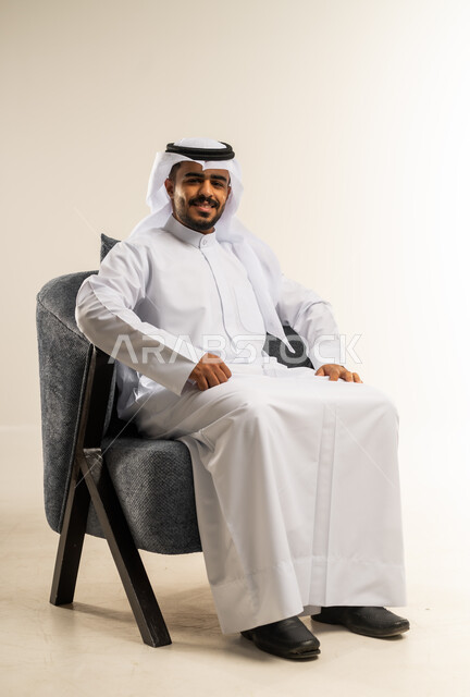 النظر الى الكاميرا بتعابير الثقة بالنفس، الاهتمام بالمظهر الخارجي والأناقة، بورتريه لرجل عربي خليجي كويتي يرتدي الثوب التقليدي يجلس على كرسي مريح، خلفية بيضاء