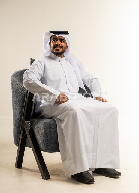 الاهتمام بالمظهر الخارجي والأناقة، مفهوم الرجولة والثقة بالنفس، بورتريه لرجل عربي خليجي كويتي يرتدي الثوب التقليدي يجلس على كرسي مريح ينظر الى شيء ما بتعابير السرور، خلفية بيضاء