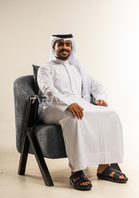 الاهتمام بالمظهر الخارجي والأناقة، مفهوم الرجولة والثقة بالنفس، بورتريه لرجل عربي خليجي كويتي يرتدي الثوب التقليدي يجلس على كرسي مريح ينظر الى الكاميرا بتعابير السرور، خلفية بيضاء