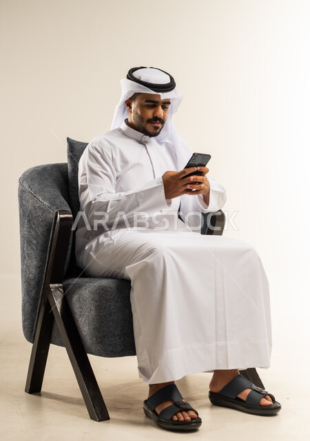 تصفح مواقع التواصل الاجتماعي, التواصل مع الأهل والأصدقاء عبر تطبيقات الجوال, بورتريه لرجل عربي خليجي كويتي يرتدي الثوب التقليدي يجلس على كرسي مريح يمسك بيده الهاتف المحمول, استخدام التقنيات الحديثة المتطورة, خلفية بيضاء