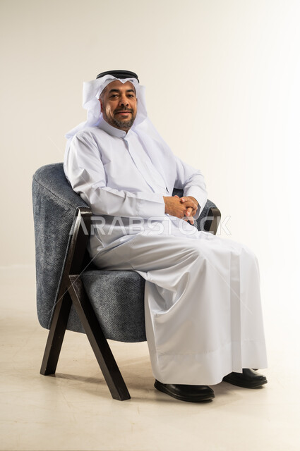 مفهوم الرجولة والثقة بالنفس، بورتريه لرجل عربي خليجي كويتي يرتدي الثوب التقليدي يجلس على كرسي مريح ينظر إلى الكاميرا بتعابير السرور، الاهتمام بالمظهر الخارجي والأناقة، خلفية بيضاء