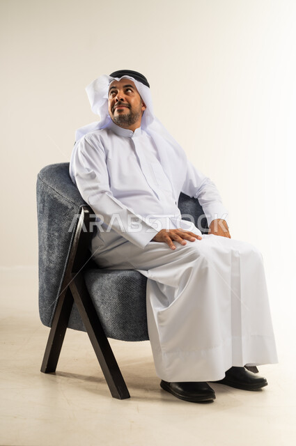 Concern for appearance and elegance, concept of masculinity and self-confidence, portrait of a Kuwaiti Gulf Arab man wearing traditional thobe sitting on a comfortable chair looking at something with expressions of pleasure, white background