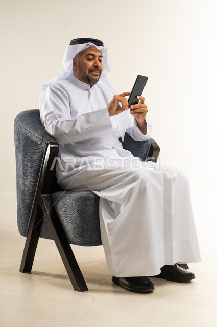 تصفح مواقع التواصل الاجتماعي, التواصل والدردشة مع الأهل والأصدقاء عبر تطبيقات الجوال, بورتريه لرجل عربي خليجي كويتي يرتدي الثوب التقليدي يجلس على كرسي مريح يستخدم الهاتف المحمول, استخدام التقنيات الحديثة المتطورة, خلفية بيضاء