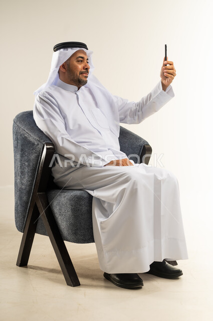 Communicating and chatting with family and friends via mobile applications, portrait of a Kuwaiti Gulf Arab man wearing traditional thobe sitting on a comfortable chair making a video call via mobile phone, using modern advanced devices and technologies, white background
