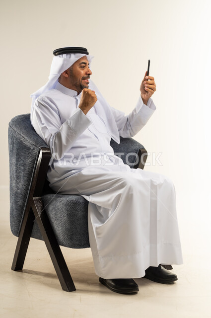 Using modern advanced devices and technologies, communicating and chatting with family and friends via mobile applications, portrait of a Kuwaiti Gulf Arab man wearing traditional dress sitting on a comfortable chair making a video call via mobile phone, white background