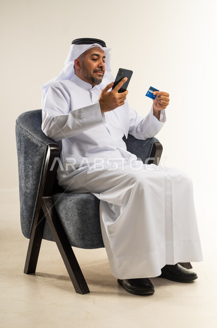 Ease of purchasing and paying by bank credit card, portrait of a Kuwaiti Gulf Arab man sitting on a chair wearing a traditional dress holding an ATM card and a mobile phone, use of modern devices and technologies, possibility of paying bills with electronic cards, white background