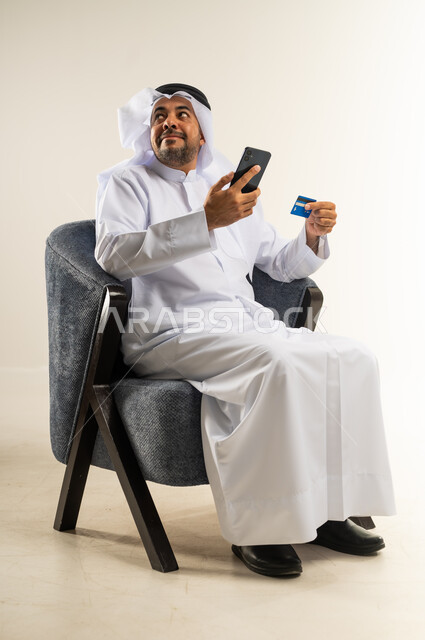 Use of modern devices and technologies, the ability to pay bills with electronic cards, ease of purchasing and paying with a bank credit card, portrait of a Kuwaiti Gulf Arab man sitting on a chair wearing a traditional dress holding an ATM card and a mobile phone, white background