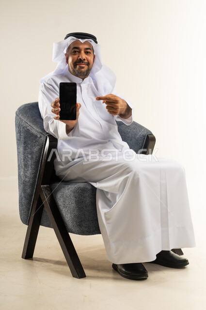 Using advanced modern devices and technologies, looking at the camera with expressions of pleasure, displaying mobile applications and programs, portrait of a Kuwaiti Gulf Arab man wearing traditional dress sitting on a comfortable chair pointing with his index finger at the empty black screen on a mobile phone, white background