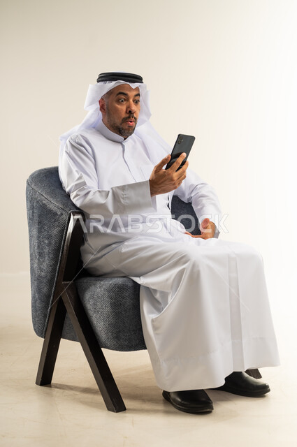 Communicating and chatting with family and friends via mobile applications, portrait of a Kuwaiti Gulf Arab man wearing traditional clothing sitting on a comfortable chair using a mobile phone looking at it with gestures of astonishment and wonder, using modern devices and technologies, white background