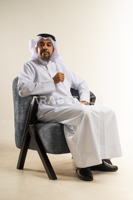 الاهتمام بالمظهر الخارجي والأناقة، مفهوم الرجولة والثقة بالنفس، بورتريه لرجل عربي خليجي كويتي يرتدي الثوب التقليدي يجلس على كرسي مريح ينظر إلى الكاميرا بتعابير السرور، خلفية بيضاء