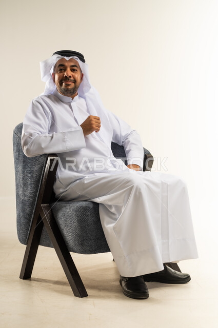 مفهوم الرجولة والثقة بالنفس، بورتريه لرجل عربي خليجي كويتي يرتدي الثوب التقليدي يجلس على كرسي مريح ينظر إلى الكاميرا بتعابير السرور، الاهتمام بالمظهر الخارجي والأناقة، خلفية بيضاء