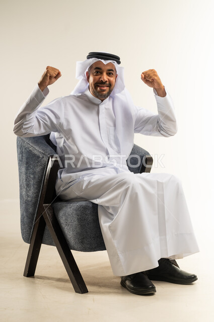 الانتصار والفوز والنجاح، مفهوم الإنجاز وتحقيق الأهداف، بورتريه لرجل عربي خليجي كويتي يرتدي الثوب التقليدي يجلس على كرسي مريح ينظر إلى الكاميرا يرفع قبضة يديه للأعلى بإيماءات الفرح والسرور, الشعور بالسعادة والقوة, خلفية بيضاء
