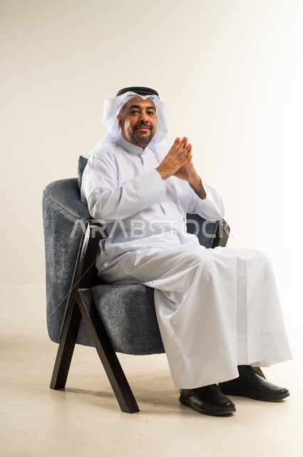 مفهوم الرجولة والثقة بالنفس، بورتريه لرجل عربي خليجي كويتي يرتدي الثوب التقليدي يجلس على كرسي مريح ينظر إلى الكاميرا بتعابير السرور، الاهتمام بالمظهر الخارجي والأناقة، خلفية بيضاء