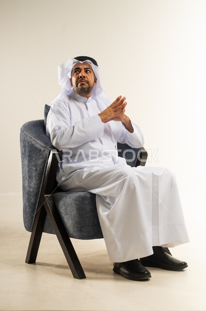 مفهوم الرجولة والثقة بالنفس، بورتريه لرجل عربي خليجي كويتي يرتدي الثوب التقليدي يجلس على كرسي مريح يضم كفيه ينظر إلى شيء ما في الأعلى، إيماءات التفكير ومحاولة تذكر أمر ما، الاهتمام بالمظهر الخارجي والأناقة، خلفية بيضاء