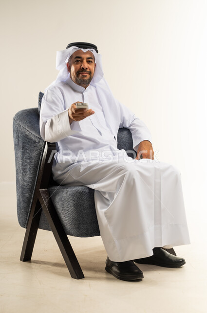 قضاء وقت ممتع بمشاهدة التلفاز، الأنشطة الترفيهية المسلية, بورتريه لرجل عربي خليجي كويتي يرتدي الثوب التقليدي يجلس على كرسي مريح يمسك بيده جهاز التحكم عن بعد, النظر إلى الكاميرا يإيماءات السرور والفرح, خلفية بيضاء