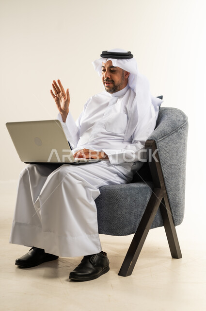 استخدام الأجهزة والتقنيات الحديثة المتطورة، عقد اجتماع عمل عبر الإنترنت، بورتريه لرجل عربي خليجي كويتي يرتدي الثوب التقليدي يجلس على كرسي مريح يرفع يده للتحية يقوم بإجراء مكالمة فيديو عبر الحاسب المحمول, إدارة شؤون الأعمال وتنظيمها، خلفية بيضاء