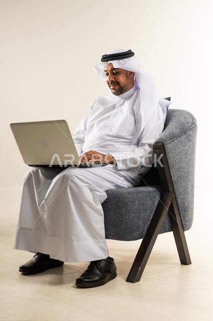 إدارة شؤون الأعمال وتنظيمها، استخدام الأجهزة والتقنيات الحديثة المتطورة، عقد اجتماع عمل عبر الإنترنت، بورتريه لرجل عربي خليجي كويتي يرتدي الثوب التقليدي يجلس على كرسي مريح يعمل عبر الحاسب المحمول, خلفية بيضاء