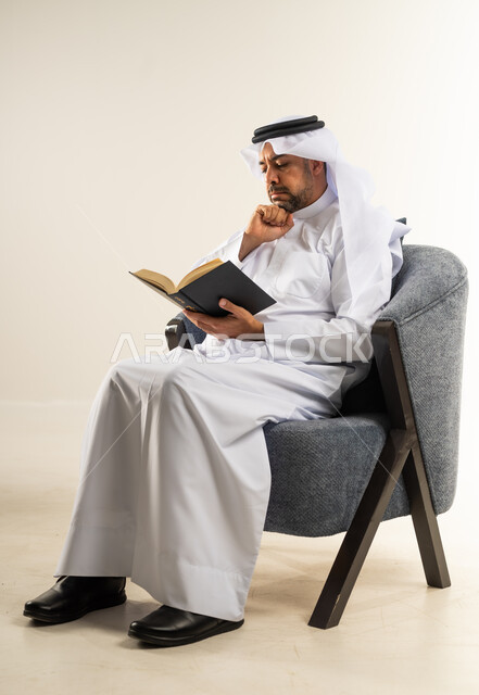 الاستمتاع بالأنشطة الترفيهية المفيدة في وقت الفراغ, قراءة القصص والروايات, بورتريه لرجل عربي خليجي كويتي يرتدي الثوب التقليدي يجلس على كرسي مريح يحمل كتاب يضع يده على ذقنه بإيماءات التفكير, ممارسة هواية المطالعة, مفهوم الثقافة والتعلم الذاتي, خلفية بيضاء