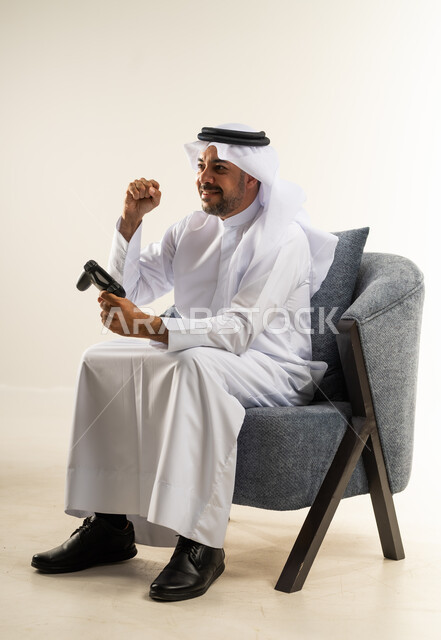 الأجهزة والتقنيات الإلكترونية الحديثة, رفع قبضة اليد للأعلى بتعابير الفوز والربح, قضاء وقت ممتع ومسلي، التفاعل والاندماج مع ألعاب الفيديو، بورتريه لرجل عربي خليجي كويتي يرتدي الثوب التقليدي يجلس على كرسي مريح يمسك بيده عصا التحكم، خلفية بيضاء