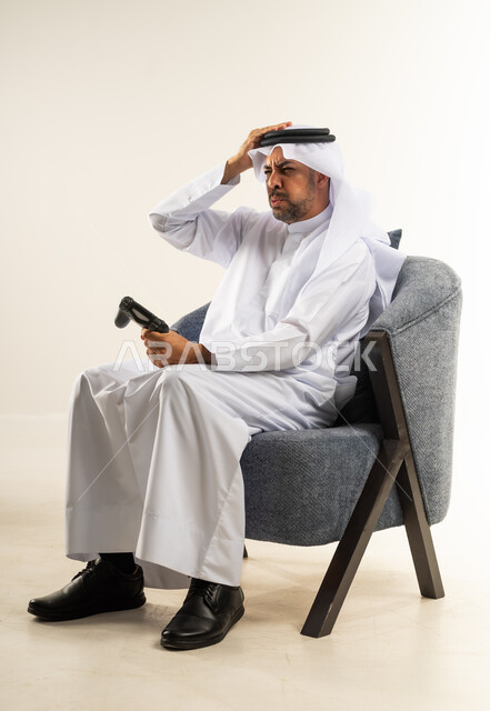 إيماءات الخسارة وخيبة الأمل، استخدام الألعاب الإلكترونية والأجهزة الحديثة، بورتريه لرجل عربي خليجي كويتي يرتدي الثوب التقليدي يمسك عصا التحكم يضع يده على رأسه بتعابير الحسرة والحزن، التسلية والترفيه بالفيديو جيمز في وقت الفراغ، خلفية بيضاء