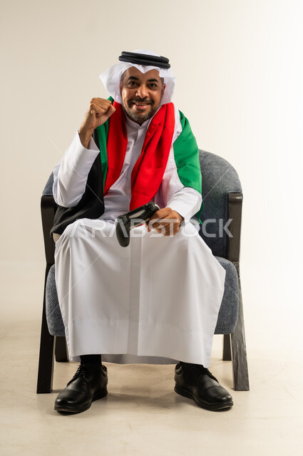 رفع قبضة اليد للأعلى بتعابير الفوز والربح, أجهزة وتقنيات إلكترونية حديثة, قضاء وقت ممتع ومسلي، التفاعل والاندماج مع ألعاب الفيديو، بورتريه لرجل عربي خليجي كويتي يرتدي الثوب التقليدي يجلس على كرسي مريح يضع علم الكويت على كتفيه يحمل بيده عصا التحكم، خلفية بيضاء